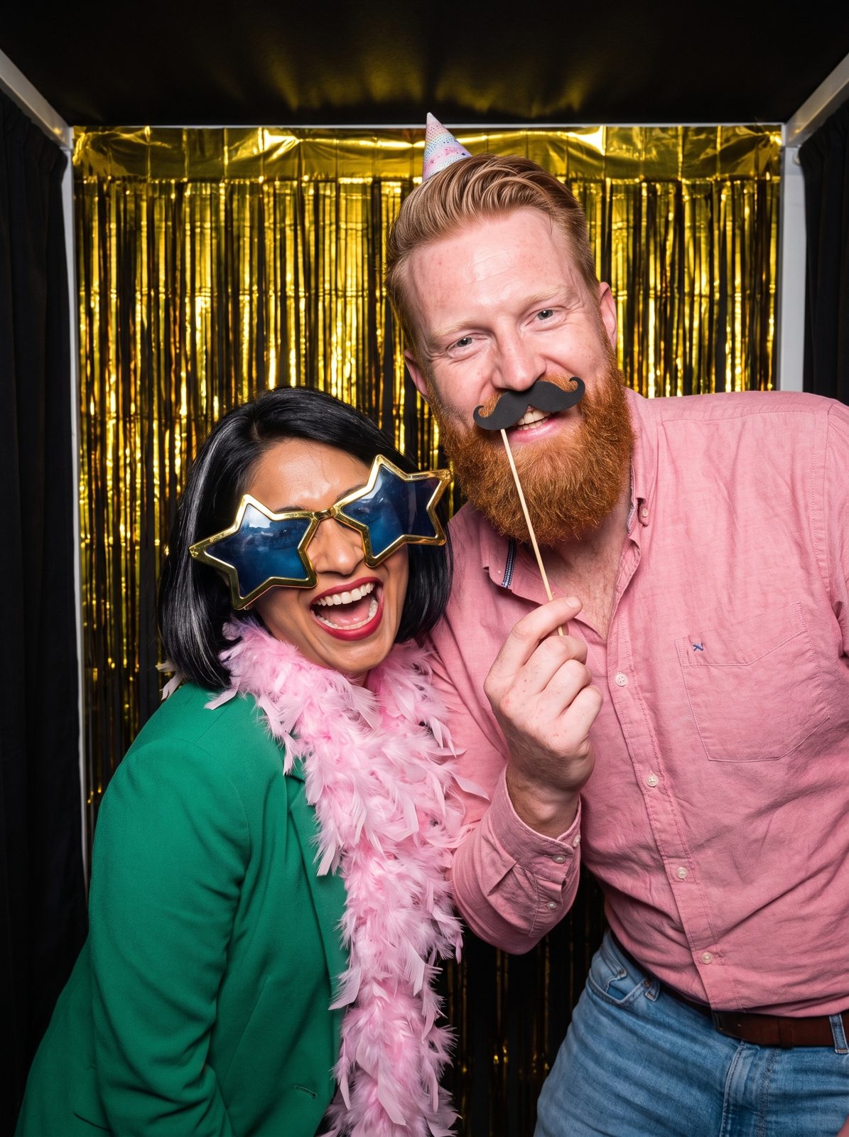 Priya in star sunglasses and a feather boa with Daan in a party hat in a photo booth.