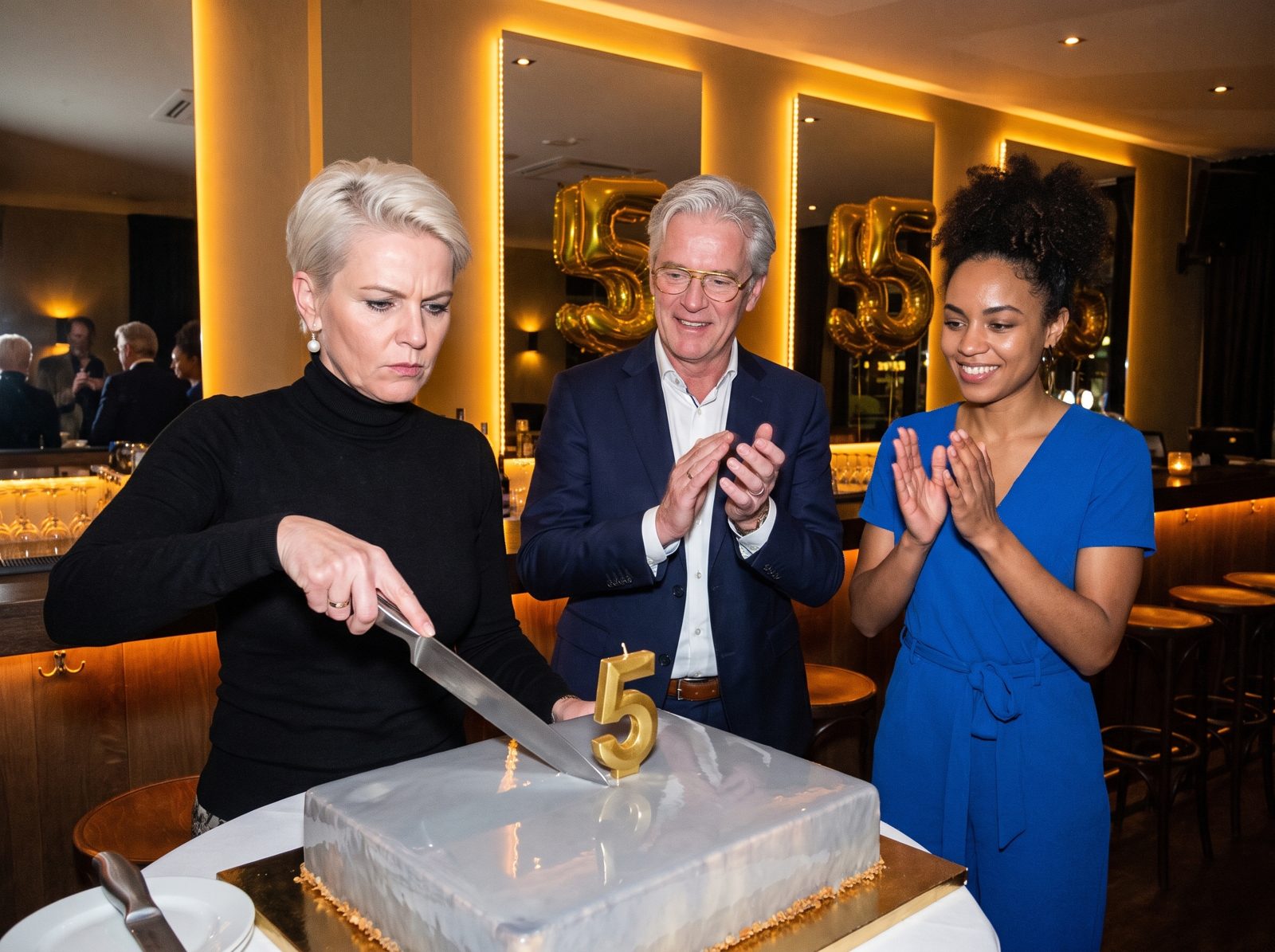 Mireille cutting a mirror-glazed cake with a golden 5 candle while Rutger and Aisha applaud.